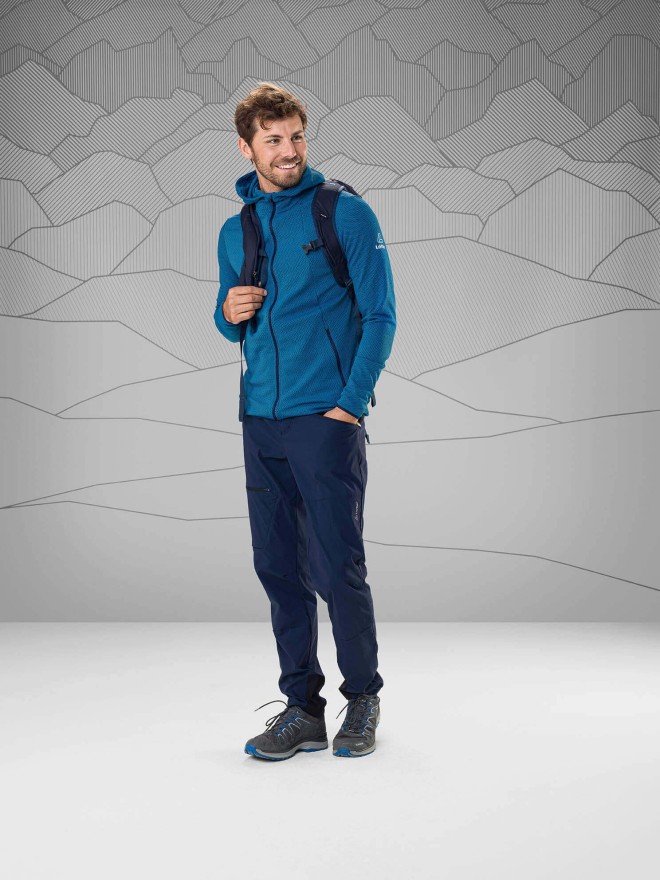 Mann mit mittelblauer Weste und dunkelblauer Wanderhose vor grauem Hintergrund, hat Rucksack umgehängt