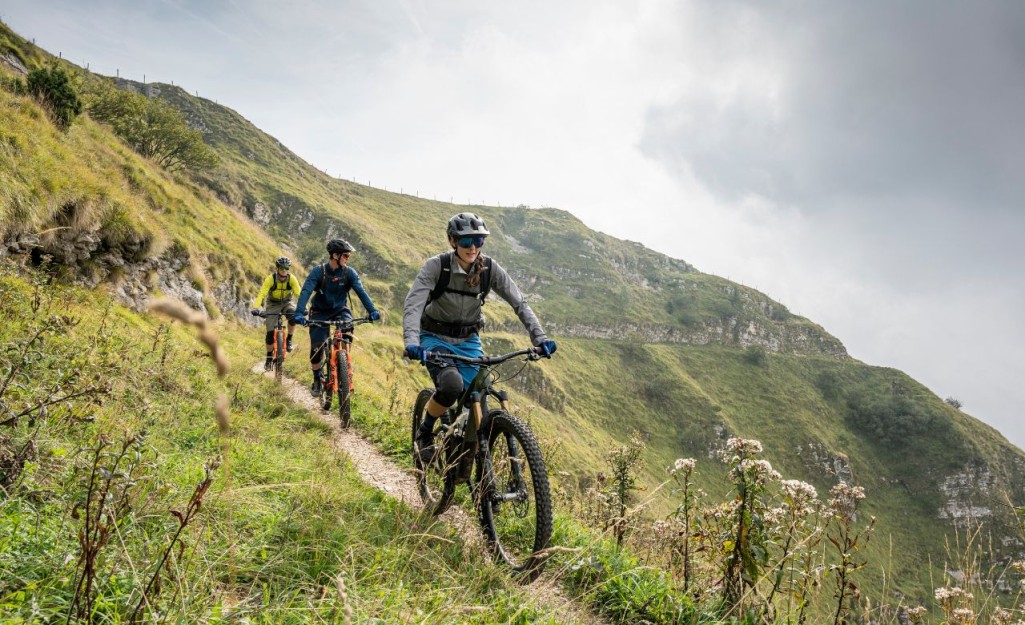 Three mountain bikers in rain jackets and bike shorts riding along a trail on a steep grassy hill.