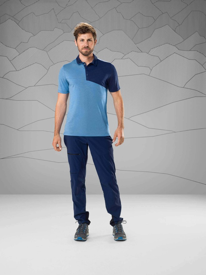 Man wearing a blue t-shirt and dark blue trekking pants standin in front of a gray background