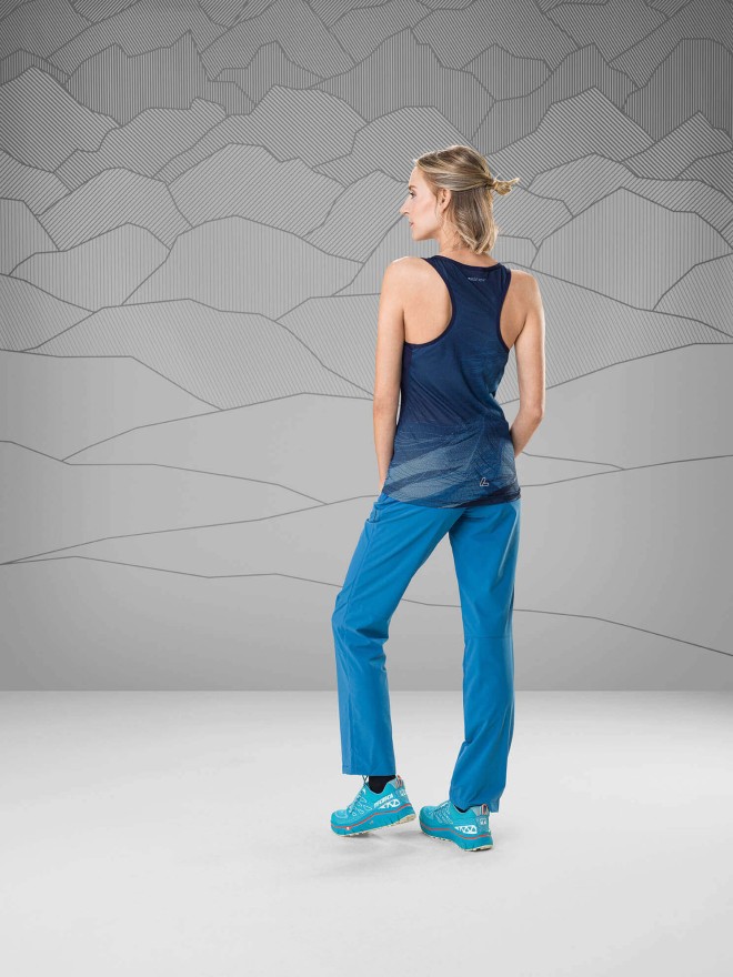 Woman wearing a dark blue racerback top and light blue trekking pants standing in front of a gray background